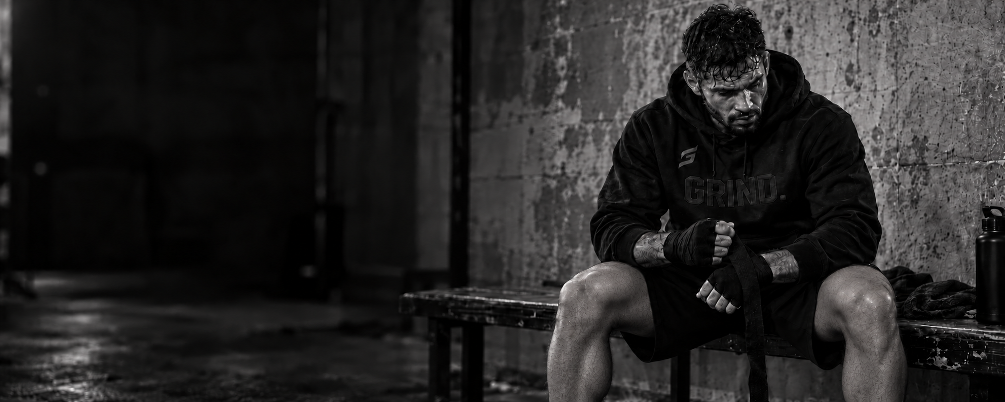 A sweat-soaked athlete in a GRIND. hoodie seated on a worn bench in a dark gym, wrapping his hands.